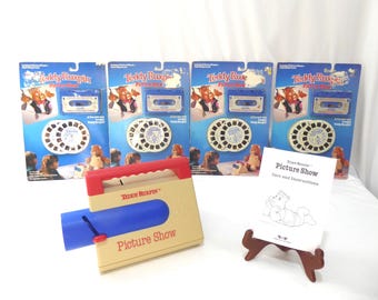 May include: A collection of vintage Teddy Ruxpin toys, including four packaged cassettes and picture reels, a tan and red picture show projector, and a care and instructions booklet. The packaging is blue with Teddy Ruxpin graphics.