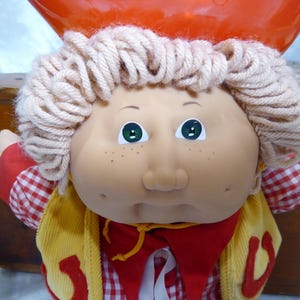 May include: A doll with light skin, freckles, and yarn hair, wearing a red and white checkered shirt under a yellow corduroy vest with red horseshoe accents. The doll has green eyes and a red neckerchief, topped with a bright orange hat.