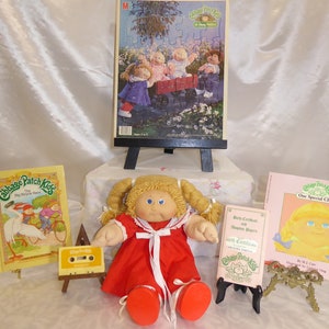 May include: A Cabbage Patch Kids doll with blonde hair and blue eyes wearing a red dress with a white ribbon. The doll is surrounded by Cabbage Patch Kids books, a cassette tape, and a puzzle.