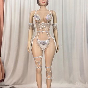 May include: A white, embellished bodysuit with sparkling accents and dangling silver chains. The costume features a halter top, arm cuffs, and leg garters, all adorned with iridescent gems. The outfit is displayed on a mannequin.