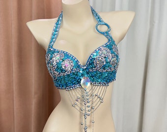 May include: A turquoise and silver sequined bra with a halter neckline and a beaded fringe. The bra has a silver ring at the top of the halter strap.
