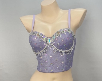Peut inclure: Un haut court de couleur lavande avec un design scintillant et iridescent. Le haut présente un corsage de style bustier avec un décolleté en cœur et des bretelles réglables. Le corsage est orné de strass et d'une bordure en franges.
