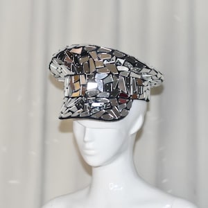 Puede incluir: Un sombrero plateado con espejos, con ala negra y corona redondeada. El sombrero está hecho de muchas piezas pequeñas y reflectantes, creando un efecto de mosaico. El sombrero se exhibe en una cabeza de maniquí blanca.