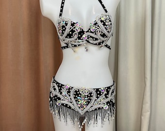 May include: Black belly dance costume with halter top and skirt. The set is adorned with iridescent rhinestones and dangling beads. The bra and skirt feature detailed patterns and a heart-shaped design. The costume is displayed on a white mannequin.