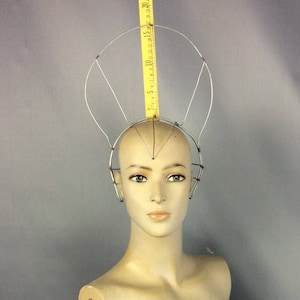 May include: A silver wire headpiece with a circular frame and three radiating spokes. The headpiece is on a mannequin head.