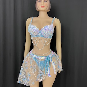 May include: A two-piece set featuring a light blue bra top covered in iridescent sequins with draped white pearl strands. The matching skirt is made of sheer mesh fabric adorned with small blue floral appliqués, iridescent sequins, and a pearl waist chain.