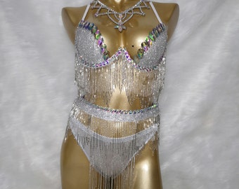 Pode incluir: Um traje de duas peças prateado e branco com franjas e pedrarias. A parte superior é um sutiã com decote halter e a parte inferior é uma saia de cintura alta.