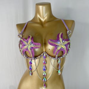 May include: A lavender and purple bra with starfish embellishments. The bra features glittery purple starfish appliqués, iridescent teardrop-shaped jewels, and silver chain detailing. The bra is designed for a costume or special occasion.
