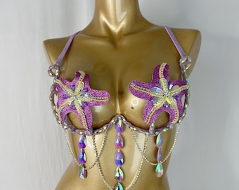May include: A lavender and purple bra with starfish embellishments. The bra features glittery purple starfish appliqués, iridescent teardrop-shaped jewels, and silver chain detailing. The bra is designed for a costume or special occasion.