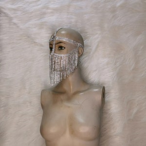 Puede incluir: Una máscara facial de pedrería plateada con una franja de pedrería. La máscara está en una cabeza de maniquí blanca.