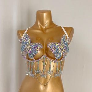 May include: A bedazzled bra with butterfly accents. The bra features iridescent blue and purple crystals arranged in a butterfly shape on the cups. Delicate beaded strands and dangling crystal bows add to the design. White straps complete the look.