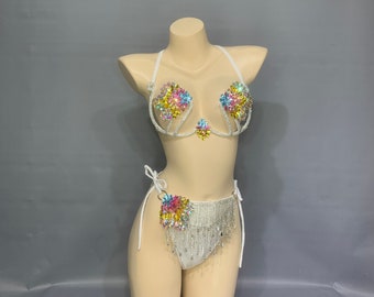 Pode incluir: Conjunto de lingerie branco com detalhes em cristal e flores coloridas. A parte de cima apresenta um design delicado de sutiã com decote profundo e a parte de baixo é uma calcinha de cintura alta estilo tanga com detalhe de franja.