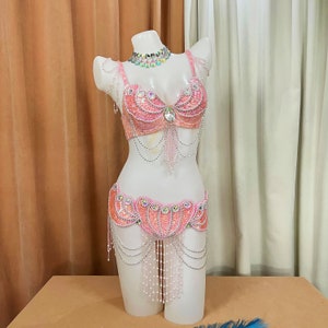 Puede incluir: Un conjunto de disfraz de danza del vientre rosa con lentejuelas sobre un maniquí. El conjunto incluye un sujetador, un cinturón y un collar, todos adornados con lentejuelas, cuentas y cadenas colgantes. El sujetador presenta una joya central y el cinturón tiene un diseño festoneado.