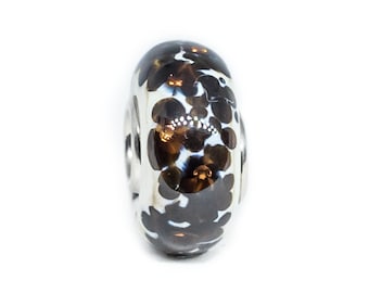 Puede incluir: Una cuenta de vidrio negra y blanca con un patrón floral. La cuenta tiene un núcleo de plata y está diseñada para usarse en un brazalete o collar.
