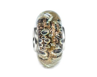Op de afbeelding: Een glazen kralen met een zilveren metalen kern. De kraal is versierd met een draaiend patroon van bruin, goud en zwart.