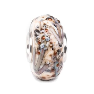 Puede incluir: Una cuenta de vidrio con una base color crema, adornada con detalles marrones, grises y azules. La cuenta presenta un patrón de formas orgánicas y pequeños detalles redondos, enmarcada por anillos de metal plateado.