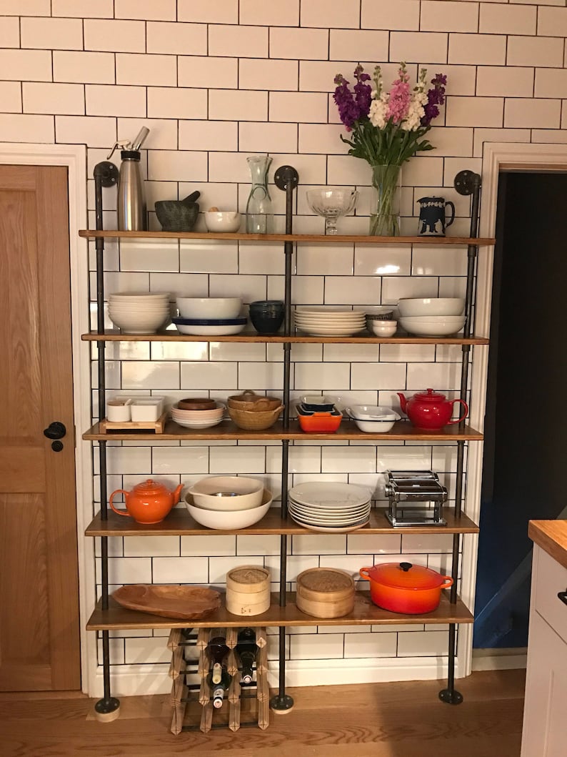 May include: A wooden shelf unit with black metal pipes, holding various kitchen items, including dishes, bowls, teapots, and a wine rack. The shelf unit is against a white tiled wall.