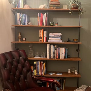 May include: A red leather armchair sits in front of a wooden bookshelf with multiple shelves. The bookshelf is filled with books and decorative items. The bookshelf is made of wood and metal pipes.