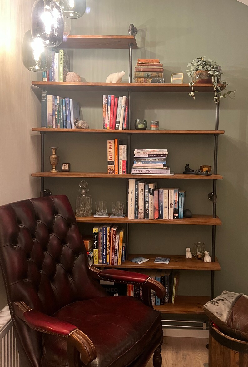 May include: A red leather armchair sits in front of a wooden bookshelf with multiple shelves. The bookshelf is filled with books and decorative items. The bookshelf is made of wood and metal pipes.