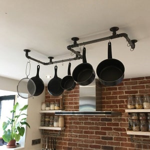 May include: A black metal pipe rack with hooks hanging from the ceiling, holding a variety of black pots and pans in a kitchen setting.