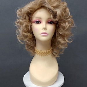 以下が含まれることがあります： ブロンドの巻き毛のウィッグと金色のビーズのチョーカーネックレスが付いたマネキンの頭。マネキンはピンクのアイシャドウと口紅をしています。頭は灰色の背景に対して白い大理石の台の上に展示されています。