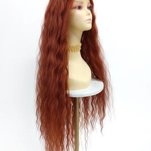 May include: A long, wavy auburn wig displayed on a mannequin head. The wig features a lace front and a natural-looking hairline. The mannequin is on a white marble-topped stand with a gold-colored metal base. The background is plain white.