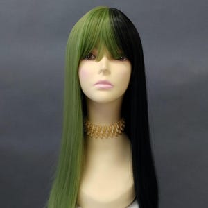 May include: A two-toned wig with a half-and-half design. One side is a vibrant green, the other is black. The wig has long, straight hair with bangs. It is displayed on a mannequin head with a gold-colored necklace.
