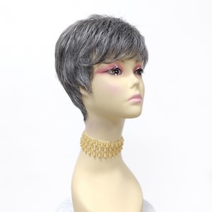 Könnte beinhalten: Eine kurze, graue Perücke mit Pony auf einem Mannequin-Kopf. Die Perücke ist in einem Pixie-Schnitt mit Seitenscheitel gestylt. Das Mannequin trägt eine goldene Halskette.