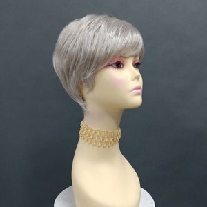 Puede incluir: Una peluca corta de color gris plateado con raya lateral y flequillo. La peluca está peinada con un corte pixie y se muestra en una cabeza de maniquí.