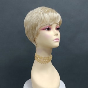 Puede incluir: Una peluca corta de color rubio con raya lateral y flequillo. La peluca está peinada con un corte pixie y se lleva en una cabeza de maniquí.