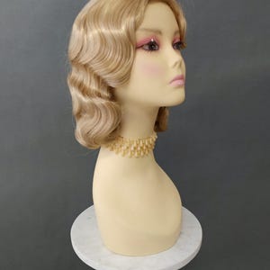 May include: A mannequin head with a blonde wig styled in waves. The mannequin has pink eye shadow and lipstick, and wears a gold beaded necklace. The head is displayed on a white marble stand.