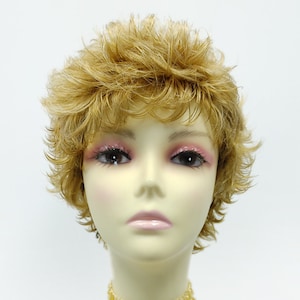 May include: A short, blonde wig with a choppy, layered style. The wig is styled on a mannequin head with a light brown skin tone. The mannequin is wearing a light brown beaded choker necklace.