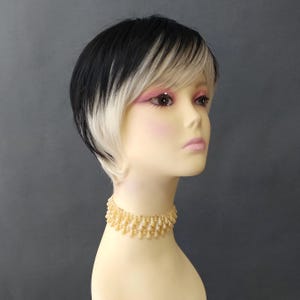 May include: A short, layered wig featuring a black crown that transitions into platinum blonde ends, styled with side-swept bangs. The wig is displayed on a mannequin head wearing a gold-toned choker necklace with dangling teardrop-shaped beads.