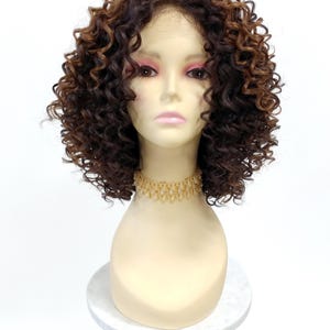 May include: A brown and auburn curly wig styled on a mannequin head. The wig features a lace front and a natural-looking hairline. A gold-colored beaded necklace is around the mannequin's neck. The mannequin head is on a white and gray marble-patterned stand.