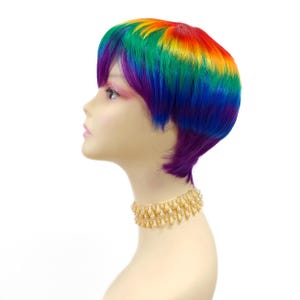 May include: A mannequin head displaying a short, rainbow-colored wig. The wig transitions from red to orange, yellow, green, blue, and purple. A gold beaded necklace adorns the mannequin's neck. The background is plain white.