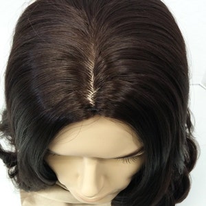 May include: A dark brown wig with a natural-looking part, styled with soft waves. The wig is displayed on a mannequin head with a neutral skin tone, against a white background. The hair appears shiny and well-maintained.