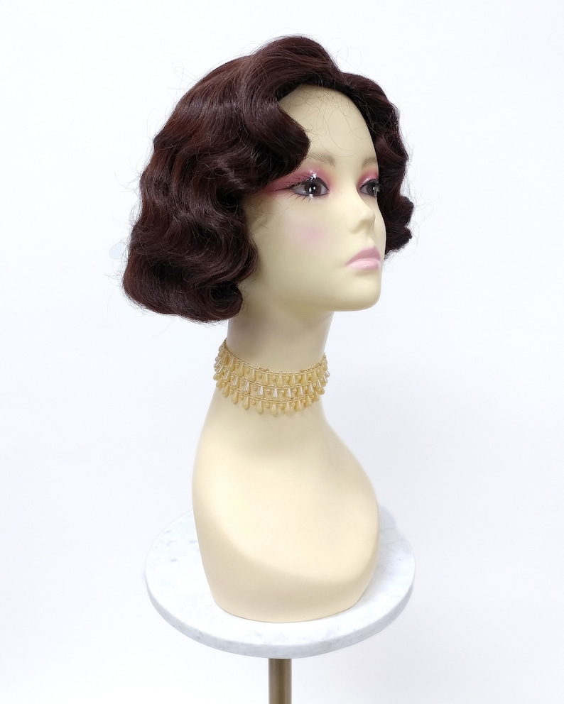 May include: A brown wig with short, curled waves styled in a vintage bob. The wig is on a mannequin head with a gold beaded choker.