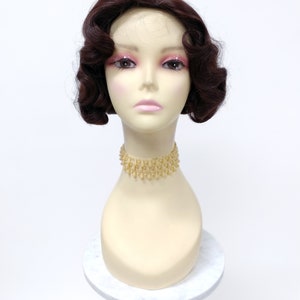 May include: A dark brown wig with short, curled waves. The wig is styled in a vintage 1920s bob haircut. The wig is on a mannequin head with a gold necklace.
