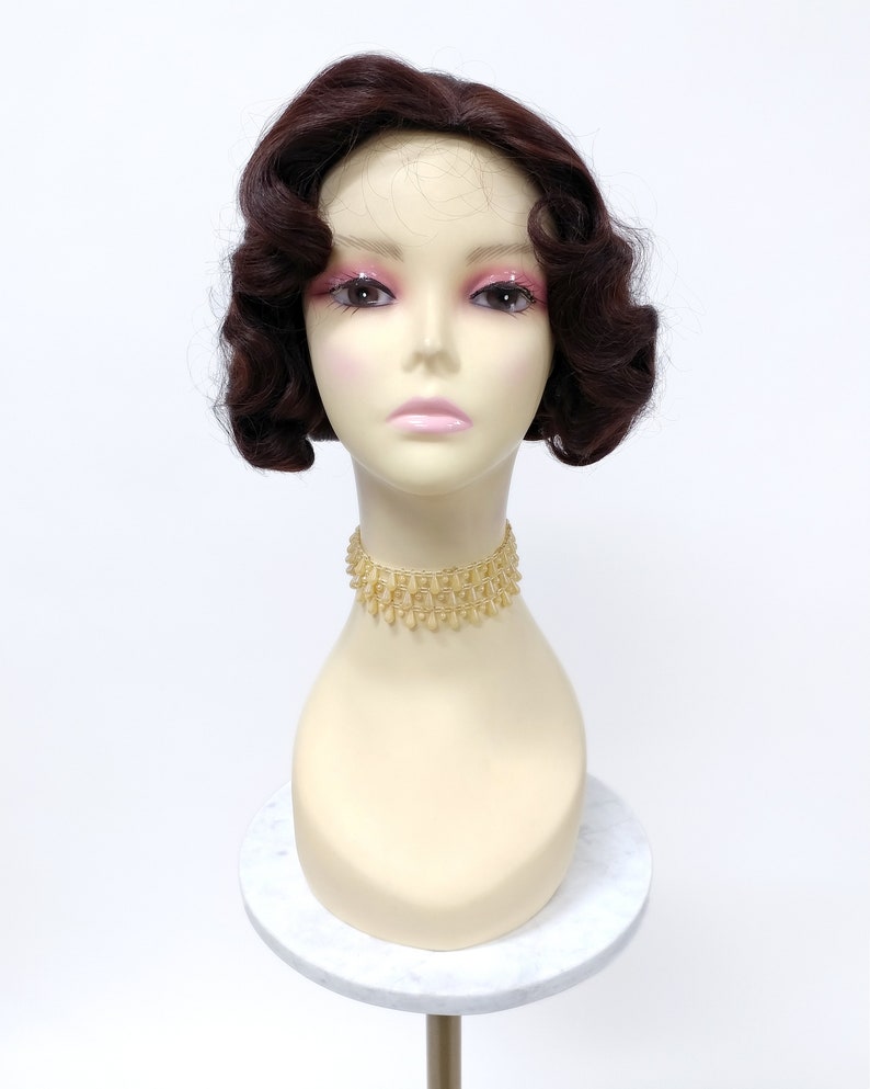May include: A dark brown wig with short, curled waves. The wig is styled in a vintage 1920s bob haircut. The wig is on a mannequin head with a gold necklace.