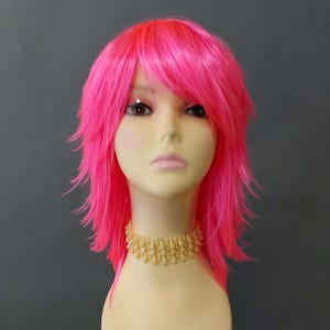 May include: A bright pink wig with a layered, shaggy style and bangs. The wig is displayed on a mannequin head, paired with a gold-toned beaded choker necklace. The wig's colour is a vivid, eye-catching shade of pink.