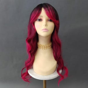 May include: A mannequin head displaying a long, wavy wig with a gradient of black to vibrant magenta. The wig features side-swept bangs and a gold-colored beaded necklace. The mannequin is on a white pedestal against a gray backdrop.