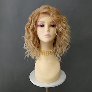 Puede incluir: Peluca rubia y rizada peinada en una cabeza de maniquí. La peluca tiene una parte delantera de encaje y está peinada con ondas sueltas. El maniquí lleva un collar dorado. La cabeza del maniquí está sobre un soporte de mármol blanco sobre un fondo gris.