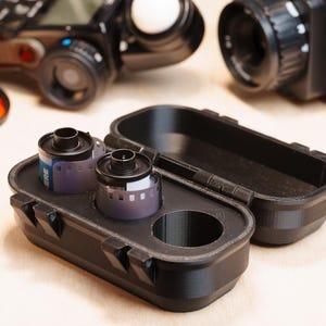 Può includere: Custodia per pellicola nera stampata in 3D con due rulli di pellicola. La custodia è aperta, rivelando le cartucce della pellicola. Una fotocamera e altre attrezzature fotografiche sono sullo sfondo. La custodia ha un coperchio incernierato e un ritaglio circolare.