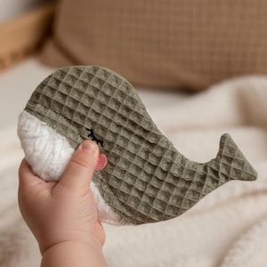 Könnte beinhalten: Ein weiches, olivgrünes Wal-förmiges Babyspielzeug mit einem strukturierten Waffelwebmuster. Der Wal hat einen weißen, flauschigen Bauch und ein kleines herzförmiges Detail in Rosa. Das Spielzeug wird in der Hand eines Kindes gehalten.