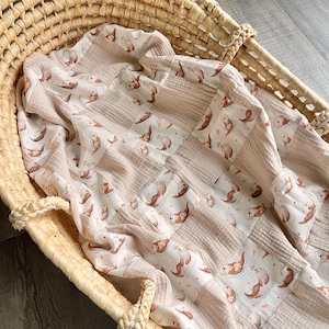 Könnte beinhalten: Eine weiche, beige Babydecke mit Patchwork-Muster, das ein sich wiederholendes Muster von braunen Ottern zeigt, die in hellblauem Wasser schwimmen. Die Decke ist gefaltet und in einem geflochtenen Korb gelegt.