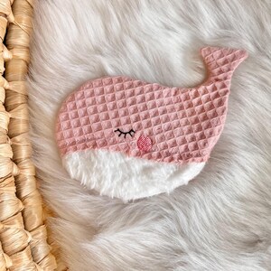 May include: A pink whale-shaped sleep mask with a textured waffle weave pattern. The whale has a white fluffy belly, embroidered eyelashes, and a small pink circle for a cheek. The mask is designed for eye coverage and relaxation.