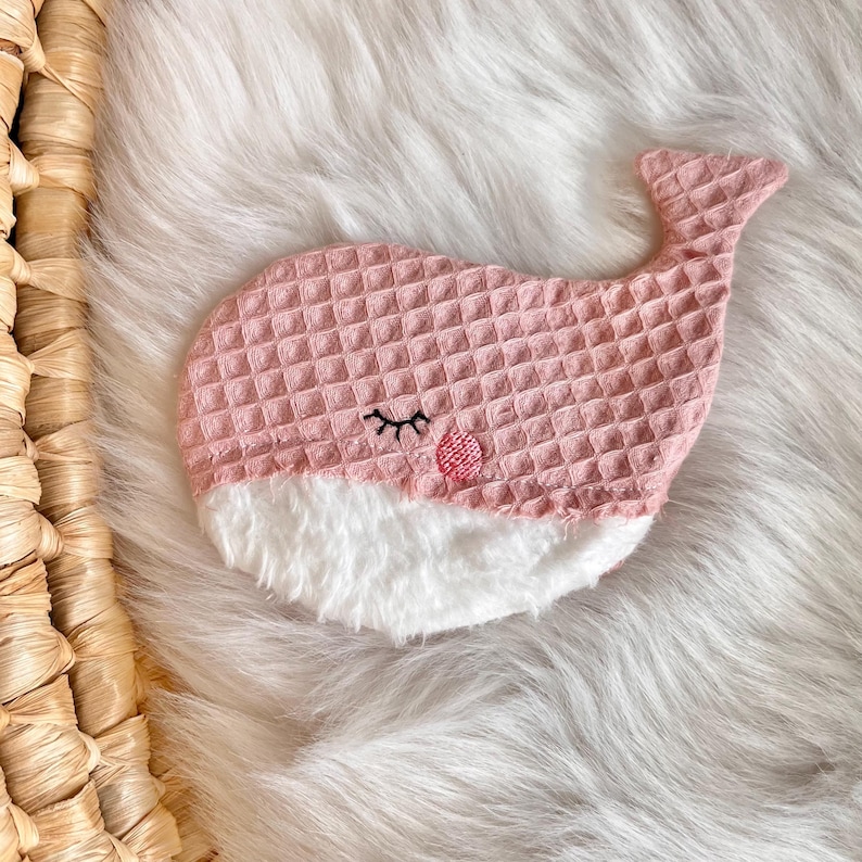 May include: A pink whale-shaped sleep mask with a textured waffle weave pattern. The whale has a white fluffy belly, embroidered eyelashes, and a small pink circle for a cheek. The mask is designed for eye coverage and relaxation.