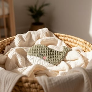 May include: A woven basket filled with a cream-colored blanket. A green whale-shaped sleep mask with a pink cheek and closed eye rests on the blanket. The basket is in a well-lit room.