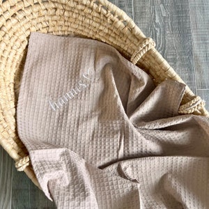May include: A light beige waffle-textured blanket featuring the embroidered name Hannes in white cursive script with a small heart outline. The blanket is draped inside a woven straw basket with a braided rim and a natural tan finish.