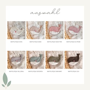 May include: A selection of eight crocheted whale-shaped baby toys in various colors, including ecru, sand, pink, and gray. Each whale has a white belly and is displayed on a textured surface.